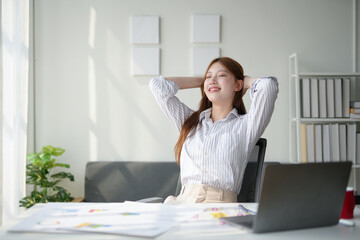 Young businesswoman relaxing at her desk in a modern office, smiling and enjoying a break from work with a laptop and documents in the background.