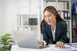 © amnaj - Businesswoman in a suit working on a laptop in a modern office environment with shelves and plants in the background.
