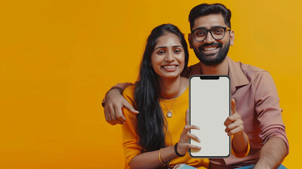 Happy indian couple sitting near big smartphone with blank white screen, demonstrating copy space for app or ad design, posing over yellow background, mockup banner