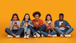 © Prostock-studio - This image features a diverse group of five young adults, all smiling and looking down at their smartphones. They are sitting cross-legged on a yellow background