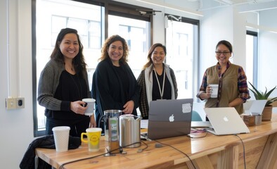 Wall Mural - Four diverse women in their thirties standing behind an office desk, smiling at the camera while holding coffee mugs and laptops