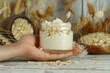 © Straxer - Oat body and facial moisturizer cream in glass jar with spikes on wooden table and hand holding spike with beige isolated background