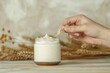 © Straxer - Oat body and facial moisturizer cream in glass jar with spikes on wooden table and hand holding spike with beige isolated background