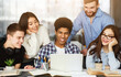 © Prostock-studio - A vibrant group of four young adults are gathered around a table filled with books, engaging in a cheerful conversation with focus on a laptop screen, doing homework together