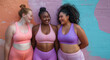 © SnapVault - Three women of diverse backgrounds stand against a vibrant wall, each wearing colorful sportswear. They are smiling and engaged in conversation, showcasing confidence, friendship, and body positivity.