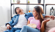 © Prostock-studio - A girl sits on a couch with a bowl of popcorn in her lap, looking annoyed as the guy beside her sleeps while watching TV. He is holding a remote control in his hand.