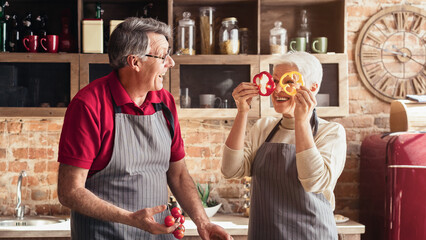 Wall Mural - Senior couple having fun on kitchen. Playful elderly woman making faces with pepper slices, free space