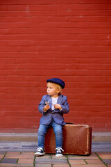 Wall Mural - Cute child, boy in vintage cloths, eating lollipop ice cream, sitting on vintage suitcase in front of a red brick wall