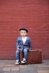 Poster - Cute child, boy in vintage cloths, eating lollipop ice cream, sitting on vintage suitcase in front of a red brick wall