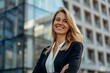 © LidiaLens - Smiling young businesswoman in a black blazer and white blouse standing confidently in front of a modern glass office building