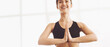 © Prostock-studio - A woman wearing a black sports bra is seen practicing yoga with her hands in the Namaste pose, while smiling in a bright studio, panorama with copy space