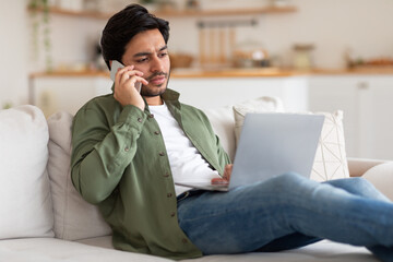 Wall Mural - A man is sitting on a white couch and talking on his cell phone. He is wearing a green shirt and blue jeans. He has a laptop on his lap. The man looks focused and serious as he speaks on the phone.