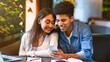 © Prostock-studio - A young black couple sitting at a table, both focused on a cell phone screen. They appear engaged with the device, possibly reading or watching something