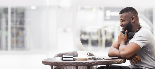 Poster - A man sits at a table in a cafe, working on his laptop while smiling. He has a cup of coffee and headphones around his neck.