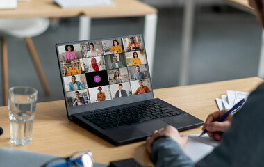Poster - A person sits at a desk in an office and participates in a video conference meeting on their laptop computer. The laptop screen displays a grid of multiple participants.