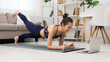 © Prostock-studio - A woman in athletic wear is doing a plank exercise on a yoga mat while watching a laptop. She is in her living room with a couch and bookshelf in the background.