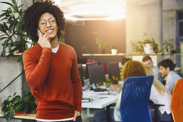Aufkleber - A cheerful young black man wearing glasses stands in the foreground, making a phone call with one hand while gesturing with the other, busy office background