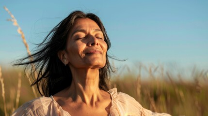 Wall Mural - A woman with closed eyes smiling and her hair blowing in the wind standing in a field with a blue sky background.