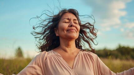 Wall Mural - Woman in pink top eyes closed smiling hair blowing in the wind standing in a field with blue sky and clouds in the background.