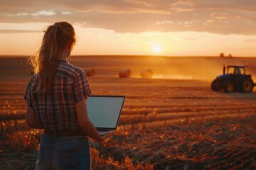 Young woman with laptop standing on field in sunset and looking at tractor baling