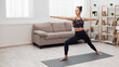 © Prostock-studio - A woman is practicing yoga in her living room, stretching her arms and legs while standing on a yoga mat.