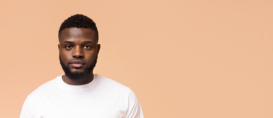 Young Black man with a short haircut and a neatly trimmed beard. He is wearing a white t-shirt and is looking directly at the camera. The background is a solid peach color, copy space