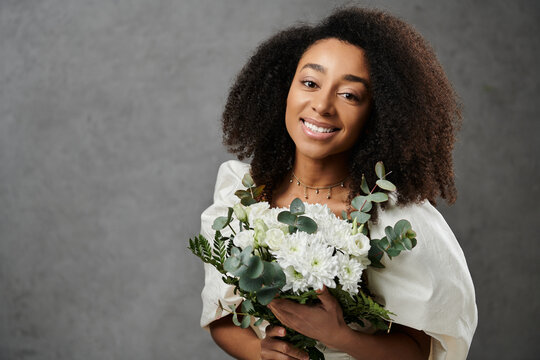A beautiful African American bride in a white wedding dress holds a bouquet of white flowers, smiling brightly against a grey backdrop.