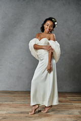 Wall Mural - A beautiful African American bride poses in a flowing white wedding dress with flowers in her hair, against a grey backdrop.