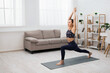 © Prostock-studio - A young woman in athletic wear practices yoga in a bright living room with white walls, hardwood floors, and a couch. She is holding a warrior one pose with her arms raised overhead.