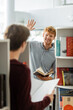 © LIGHTFIELD STUDIOS - A man stands in front of a bookshelf and his friend, holding a book.