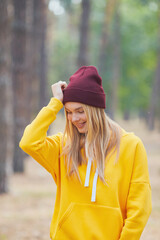 Wall Mural - Blue-eyed blonde in a yellow hoodie and burgundy hat in a pine forest. Portrait of a joyful young woman enjoying in autumn park.