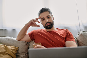 a bearded man in casual attire works remotely from home on a couch, using a laptop.