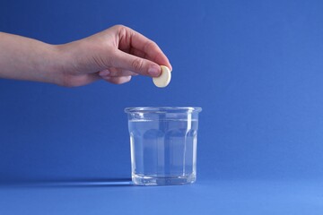 Wall Mural - Woman putting effervescent pill into glass of water on blue background, closeup