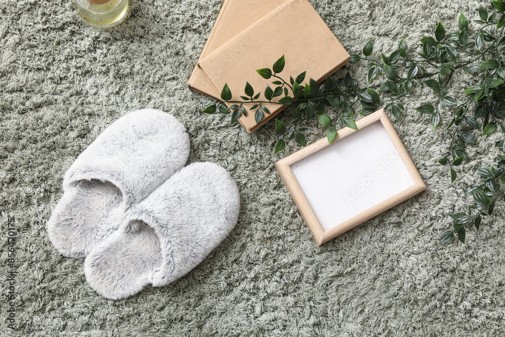 Soft slippers with books, blank frame and plant branches on green carpet, top view