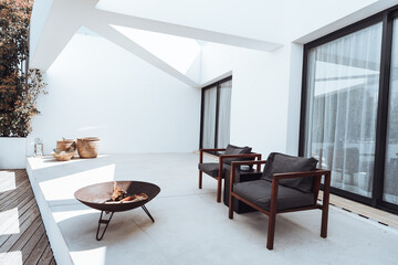 A modern outdoor patio with minimalist design featuring two wooden chairs with dark cushions, a metal fire pit, and potted plants against white walls. Large glass doors with curtains inside