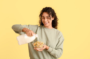 Wall Mural - Emotional young woman pouring milk into bowl of tasty cereal rings on color background