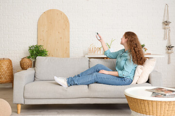 Canvas Print - Young woman with air conditioner remote control sitting on sofa in living room