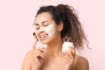 Wall Mural - Young African-American woman with jar of facial mask and brush on pink background