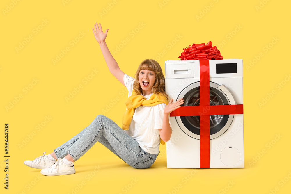 Happy young woman with gift washing machine on yellow background