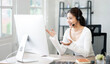 © NAMPIX - Beautiful asian woman with headset doing video conference via desktop computer sitting at home office.