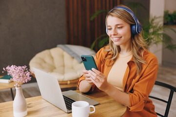 Wall Mural - Young IT woman wear orange shirt headphones hold mobile cell phone using laptop pc computer listen music sit alone at table in coffee shop cafe indoor work or study. Freelance office business concept.