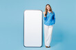 © ViDi Studio - Full body young woman wear shirt casual clothes point thumb finger on big huge blank screen mobile cell phone smartphone with copy space area isolated on plain light blue background Lifestyle concept