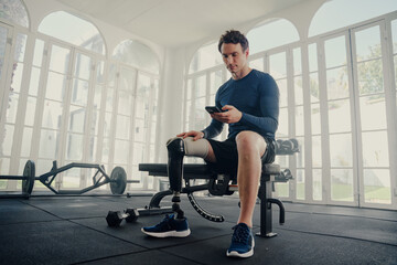 Wall Mural - Young caucasian man with prosthetic leg sitting using mobile phone on bench press at the gym