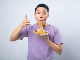 Young Asian man in a lavender t-shirt enthusiastically eating instant noodles from a bowl with a fork. Studio shot on a plain background, highlighting his joyful expression and casual fashion