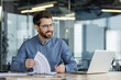 © Tetiana - A smiling young man sits in the office at a table in front of a laptop, makes notes in documents, works with data