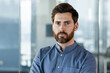 © Tetiana - Close-up portrait of a young serious businessman in a blue shirt standing inside the office and looking confidently at the camera