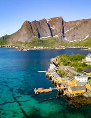 Wall Mural - Aerial view of a small Norwegian village with colorful houses on stilts along the turquoise waters of a fjord. Mountains rise in the background, providing a picturesque backdrop. Sakrisoy, Lofoten