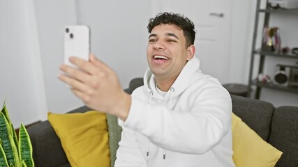 Wall Mural - A smiling young man with curly hair takes a selfie indoors on his smartphone, comfortably lounging in a neutral-hued room.