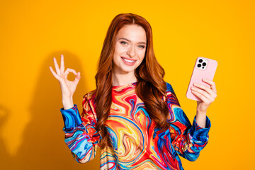 Wall Mural - Photo of satisfied girl with foxy hair dressed print shirt hold smartphone showing okey good work isolated on yellow color background