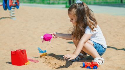 Pretty child girl playing in sand on outdoor playground. Beautiful baby in jeans shorts having fun on sunny warm summer day. Child with colorful sand toys. Healthy active baby outdoors plays games